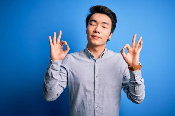 孤立した青の背景の上に立ってカジュアルなシャツを着て若いハンサムな中国人男性がリラックスし 目を閉じて指で瞑想のジェスチャーをして笑顔 ヨガのコンセプト — ストック写真