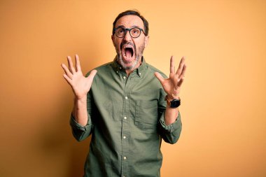 Orta yaşlı, yeşil tişört giyen, gözlüklü, izole edilmiş sarı arka planda çılgın deli bağıran, agresif bir ifade ve kollarla bağıran orta yaşlı bir adam. Hayal kırıklığı konsepti.