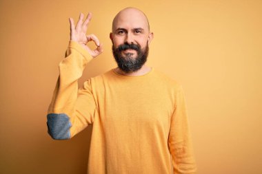 Sarı arka planda dikilmiş sakallı yakışıklı, kel adam el ve parmaklarıyla tamam işareti yaparken gülümsüyor. Başarılı ifade.