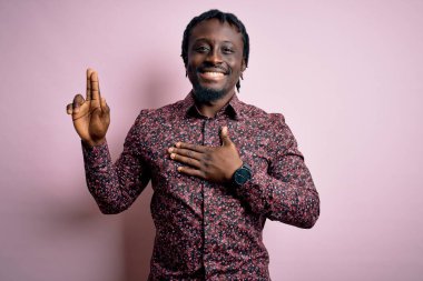 Genç, yakışıklı, Afro-Amerikalı bir adam pembe arka planda dikilmiş, gülümsüyor, yemin ediyor ve bağlılık yemini ediyor.