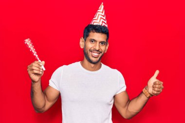 Doğum günü şapkası takan genç Latin adam mutlu ve pozitif gülümsüyor, başparmağıyla mükemmel ve onay işareti yapıyor. 