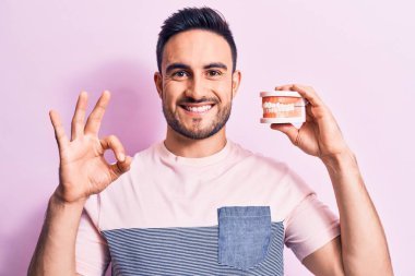 Sakallı, takma dişli, pembe arka planda genç yakışıklı adam parmaklarıyla işaret veriyor, gülümseyerek mükemmel bir sembol oluşturuyor.