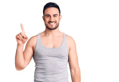 Mayo ve kolsuz tişört giyen genç yakışıklı bir numaralı parmağını gösterip onu gösterirken kendinden emin ve mutlu bir şekilde gülümsüyor.. 