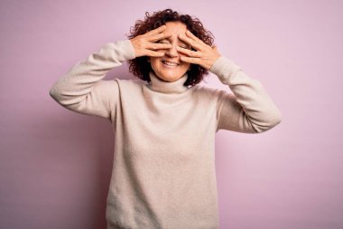 Orta yaşlı güzel kıvırcık saçlı kadın pembe arka plan üzerine balıkçı yaka kazak giyip gözleri kapayarak neşeli ve komik gülümsüyor. Kör kavram.