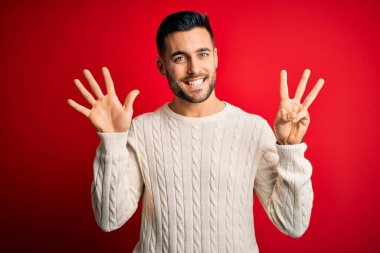 İzole edilmiş kırmızı arka plan üzerinde duran, rahat beyaz süveter giyen genç yakışıklı adam 8 numaralı parmaklarıyla işaret ederken kendinden emin ve mutlu gülümsüyor..