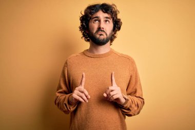 Sakallı, sıradan bir süveter giyen, sarı arka planda dikilen üzgün ve üzgün görünen, parmaklarıyla yönü gösteren, mutsuz ve depresif genç bir adam..