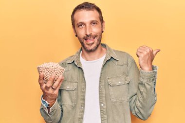 Genç yakışıklı adam, elinde nohut kasesi, başparmağını yana kaldırmış, ağzı açık gülümsüyor. 