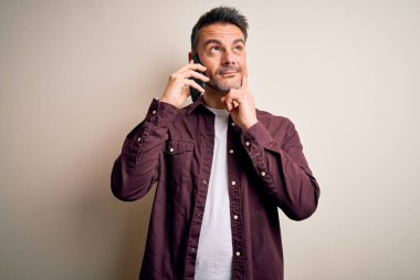 Genç yakışıklı bir adam beyaz arka planda akıllı telefondan konuşuyor. Ciddi bir yüz ifadesi var. Soru sormayı düşünüyor.