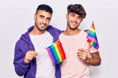 Genç eşcinsel çift ellerinde gökkuşağı lgbtq bayrağı tutuyorlar pozitif ve mutlu görünüyorlar ve gülümsüyorlar dişlerini gösteren bir gülümsemeyle 