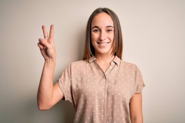 İzole edilmiş beyaz arka plan üzerinde duran rahat tişört giyen genç ve güzel bir kadın iki numaralı parmaklarıyla işaret ederken kendinden emin ve mutlu bir şekilde gülümsüyor..
