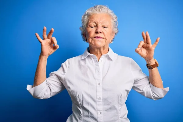 年长的漂亮女人穿着雅致的衬衫 站在孤立的蓝色背景上放松 闭上双眼微笑 用手指做冥想动作 瑜伽概念 — 图库照片