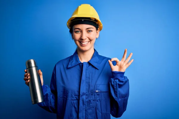 Young beautiful brunette worker woman wearing safety helmet holding thermo with water doing ok sign with fingers, excellent symbol