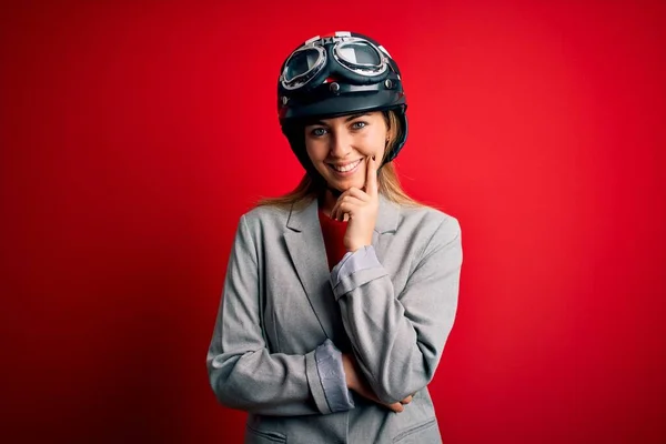 若い美しいブロンドのオートバイの女性は 交差腕と手で笑顔でカメラを見て自信を持って見える赤い背景の上にオートバイのヘルメットを着てあごに提起 肯定的な考え — ストック写真