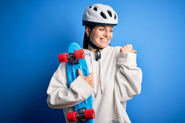 年轻美丽的红头发滑板女运动员 戴着安全帽 手握滑板指向一侧 带着大拇指朝侧看 笑容满面 — 图库照片