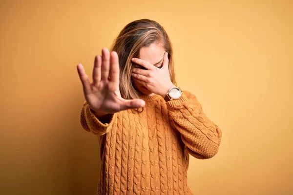 Young beautiful blonde woman wearing casual sweater standing over yellow background covering eyes with hands and doing stop gesture with sad and fear expression. Embarrassed and negative concept.