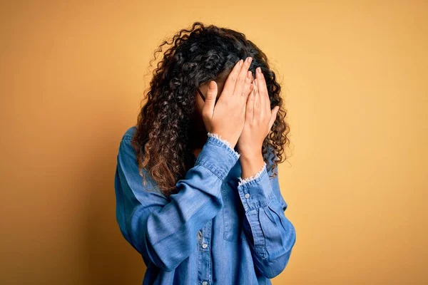 Beautiful woman with curly hair wearing casual denim shirt and glasses over yellow background with sad expression covering face with hands while crying. Depression concept.