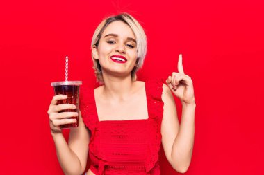 Genç güzel sarışın kadın bir bardak kola içiyor. Bir fikir ya da soruyla şaşırmış. Mutlu yüzle işaret ediyor. 