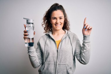 Kıvırcık saçlı, spor yapan beyaz arka planda bir şişe su içen güzel bir kadın bir fikir ya da soru işareti ile şaşırdı.