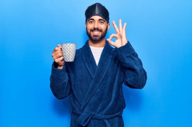 Uyku maskesi takan ve sabahlık giyen genç İspanyol adam kahve içiyor. Parmaklarıyla imza atıyor, gülümsüyor ve mükemmel bir sembol oluşturuyor. 