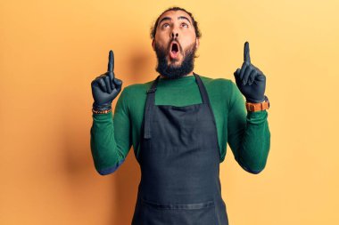 Berber önlüğü giymiş genç Arap adam şaşırmış ve şaşırmış bir şekilde yukarı bakıyor ve parmaklarıyla işaret ediyor ve kollarını kaldırıyordu.. 