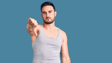 Mayo ve kolsuz tişört giyen genç yakışıklı adam mutsuz görünüyor ve reddediliyor ve baş parmakları aşağıda hareket ediyor. kötü ifade. 