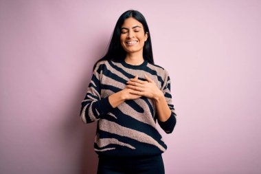 Pembe arka planda hayvan desenli süveter giyen, elleri göğsünde, gözleri kapalı ve yüzünde minnet dolu bir jest olan genç ve güzel bir İspanyol kadın. Sağlık kavramı.