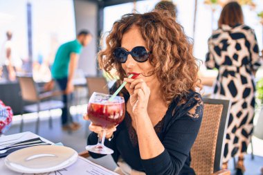 Orta yaşlı, güzel, güneş gözlüğü takan esmer kadın. Teras restoranında oturmuş bir bardak sangria içiyorum.