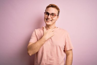 Genç, yakışıklı, günlük tişört giyen, pembe arka planda duran neşeli, yüzünde gülümsemesi olan mutlu ve doğal ifadeyle elini ve parmağını kaldıran bir adam.