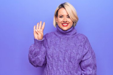 Güzel sarışın ve cüsseli bir kadın mor arka planda balıkçı yaka kazak giyiyor ve parmaklarını dört numarayı gösterirken kendinden emin ve mutlu bir şekilde gülümsüyor..
