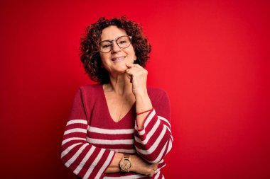 Orta yaşlı güzel kıvırcık saçlı kadın kırmızı arka planda sıradan çizgili süveter giyiyor kameraya kendinden emin bakıyor kollarını kavuşturup gülümseyerek ve çenesini kaldırarak. Olumlu düşünüyorum..