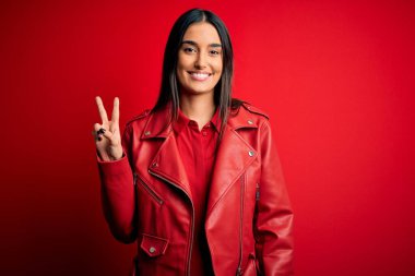 Genç, güzel, esmer, günlük ceket giyen, izole edilmiş kırmızı arka planda dikilen ve kameraya gülümseyen, zafer işareti yapan bir kadın. İki numara..