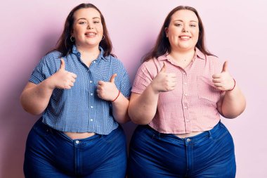 Genç ve cüsseli ikizler gündelik kıyafet giyiyorlar. Başarı işareti yapıyorlar. Elleriyle olumlu jest yapıyorlar. Neşeli ifade ve kazanan jest. 