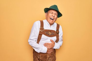 Kıdemli gri saçlı adam sarı arka planda geleneksel Alman Ekim Festivali kıyafeti giyiyor. Gülümsüyor ve kahkahalarla gülüyor..
