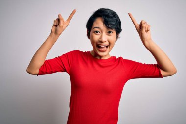 Genç, güzel Asyalı bir kız, günlük kırmızı tişört giyiyor, beyaz arka plan üzerinde duruyor. Şaşırmış, şaşırmış ve parmaklarıyla işaret ediyor ve kollarını kaldırıyor..