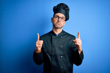 Genç, yakışıklı aşçı üniformalı ve şapkalı, izole edilmiş mavi arka planda üzgün ve üzgün görünüyor, parmaklarıyla yönü gösteriyor, mutsuz ve depresif..