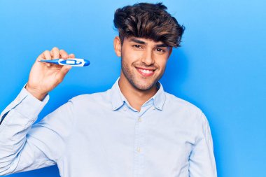 Genç İspanyol adam termometreyi tutuyor. Pozitif ve mutlu görünüyor. Durup gülümsüyor ve kendine güvenen bir gülümsemeyle dişlerini gösteriyor. 