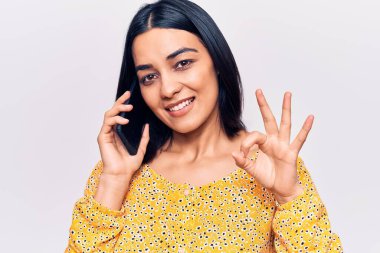 Genç, güzel, Latin bir kadın akıllı telefonda konuşuyor parmaklarıyla imza atıyor, gülümsüyor ve mükemmel bir sembol oluşturuyor. 