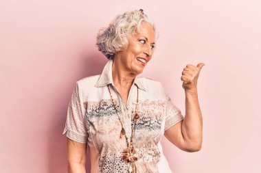 Yaşlı, gri saçlı, günlük kıyafetler giyen, gülümseyen, gülümseyen ve baş parmağıyla yüzünü gösteren bir kadın.. 