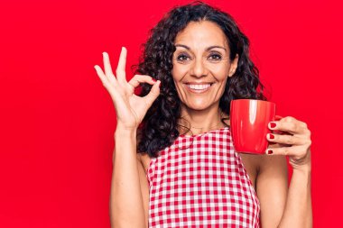 Orta yaşlı güzel bir kadın bir kupa kahve içiyor parmaklarıyla imza atıyor, gülümseyerek mükemmel bir sembol oluşturuyor. 