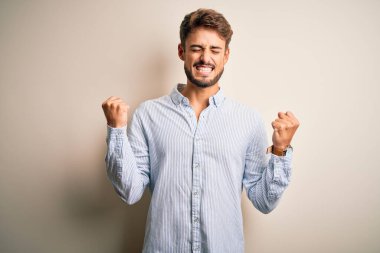 Beyaz arka plan üzerinde duran çizgili tişörtlü, sakallı yakışıklı genç adam çok mutlu ve heyecanlı bir şekilde kollarını kaldırıp, gülümseyerek ve başarı için çığlık atarak zafer hareketi yapıyor. Kutlama konsepti.