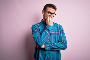 Rahat gömlekli ve gözlüklü genç bir adam pembe arka planda duruyor gergin ve gergin görünüyor ağızları tırnak yiyor. Endişe sorunu.