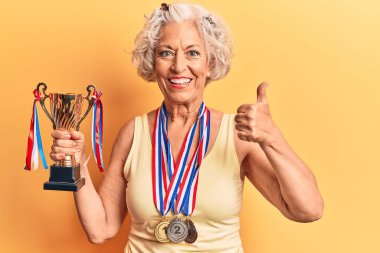 Kıdemli gri saçlı kadın elinde şampiyonluk kupası tutuyor mutlu ve pozitif gülümsüyor, başparmağıyla mükemmel ve onay işareti yapıyor. 