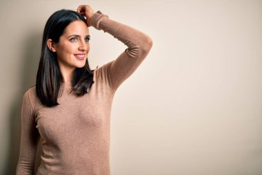 Young brunette woman with blue eyes wearing casual sweater over isolated white background smiling confident touching hair with hand up gesture, posing attractive and fashionable