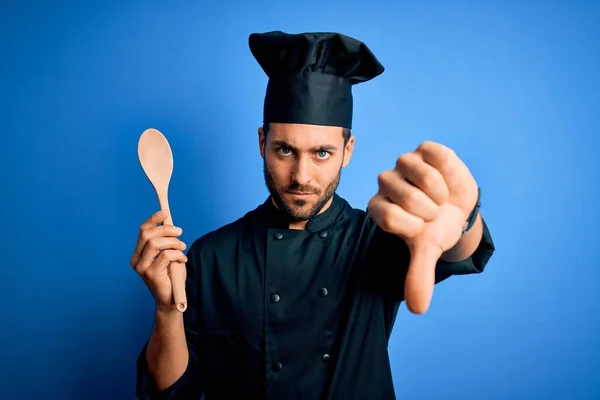 年轻的大胡子厨师 身穿制服 头戴木制勺子 头戴蓝色背景 满脸怒容 不喜欢大拇指朝下的负面标志 拒绝的观念 — 图库照片