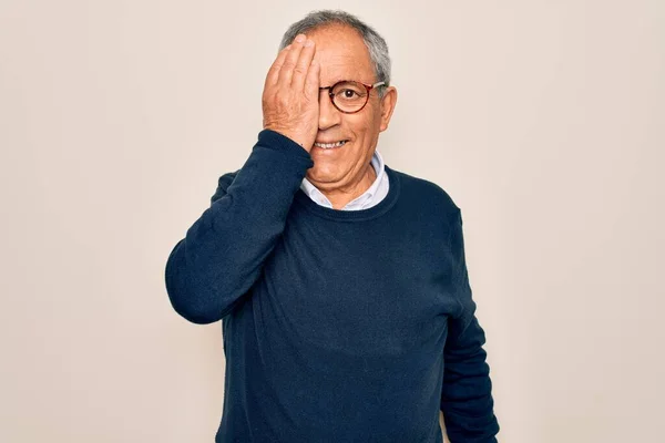 Senior handsome grey-haired man wearing sweater and glasses over isolated white background covering one eye with hand, confident smile on face and surprise emotion.