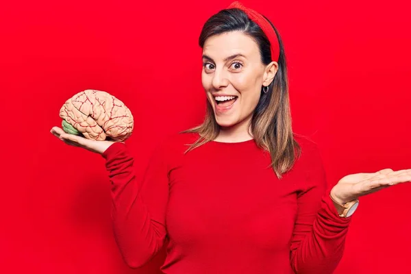 Young beautiful woman holding brain celebrating achievement with happy smile and winner expression with raised hand