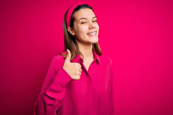 若い美しいブルネットの女の子は 手で幸せな親指アップジェスチャーを行う孤立したピンクの背景の上に立ってカジュアルなシャツを着ています 成功を示すカメラを見て表現を承認する — ストック写真
