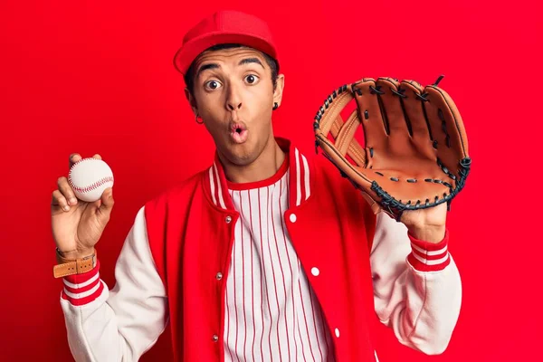 Young african amercian man wearing baseball uniform holding golve and ball scared and amazed with open mouth for surprise, disbelief face