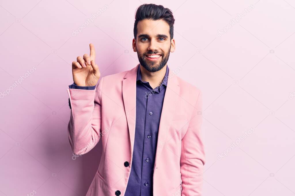 Young handsome man with beard wearing elegant jacket showing and pointing up with finger number one while smiling confident and happy. 