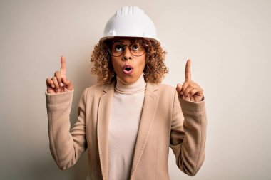 Genç Afro-Amerikan kıvırcık mimar kadın güvenlik kaskı ve gözlük takıyor hayret ediyor, şaşırıyor ve parmaklarıyla işaret ediyor ve kollarını kaldırıyor..
